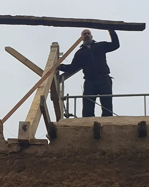 Julien-Pierre Hubert sur un chantier, guidant une pièce de charpente en cours de levage au-dessus d’un mur en terre